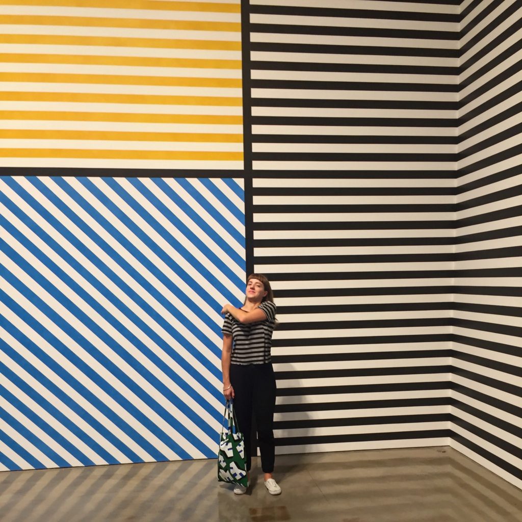 Joelle Te Paske stands in front of a wall drawing by Sol LeWitt, with repeating lines in yellow, blue, and black